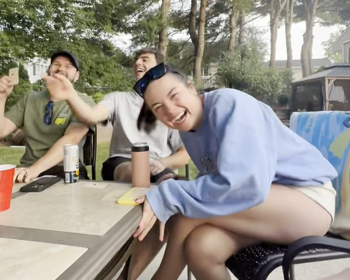 Group of people laughing sitting around a table outdoors under a blue umbrella with trees in the background